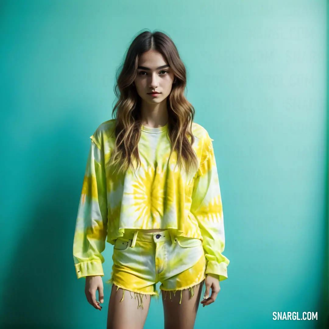 Woman in yellow shorts and a tie dye shirt standing in front of a blue wall with her hands on her hips