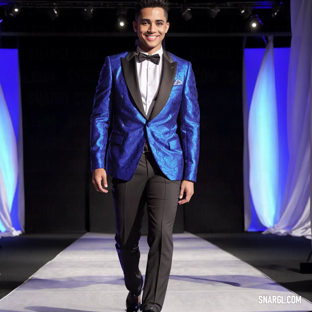 Man in a blue tuxedo walks down a runway at a fashion show wearing a black bow tie