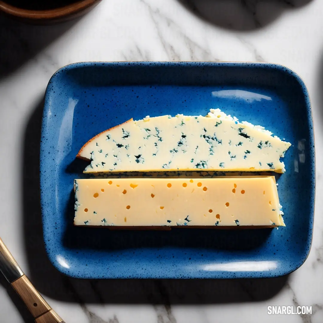 Blue plate with a piece of cheese on it and a knife on the side of it on a marble table. Color Cobalt.