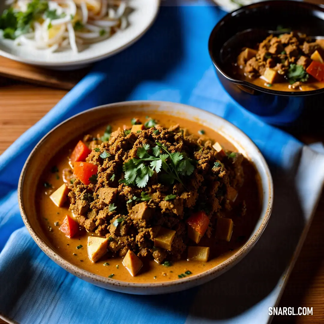 Bowl of food on a table with a blue napkin and a bowl of soup on the side of the table. Example of #B8860B color.