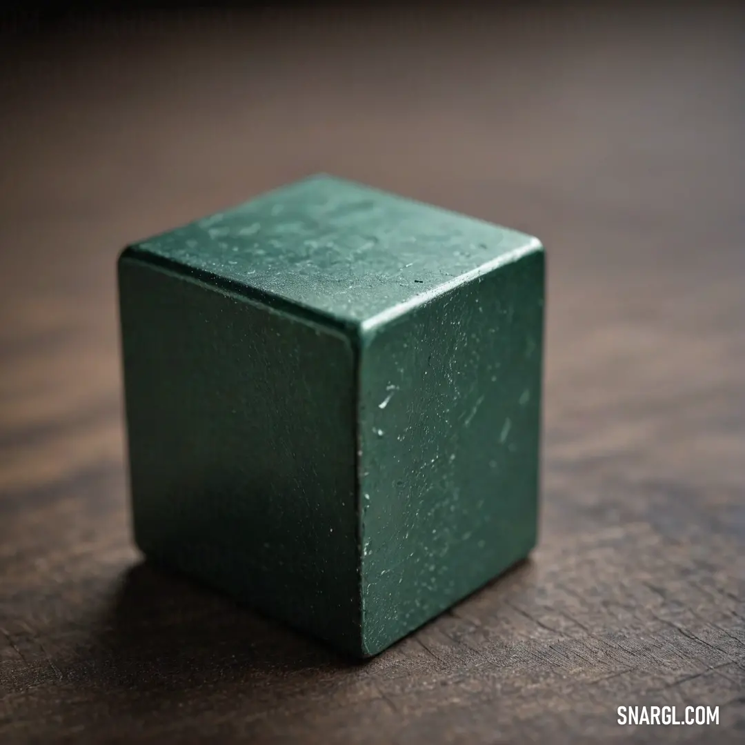 Green cube on top of a wooden table next to a black object with a white spot on it. Example of Dark green color.