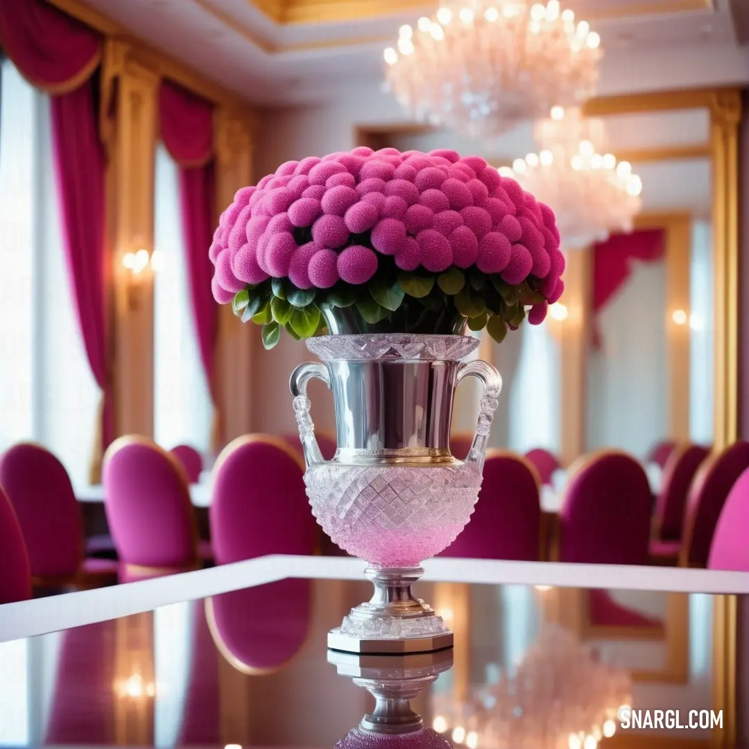 Vase with flowers on a table in a room with pink chairs and chandeliers in the background. Color #872657.