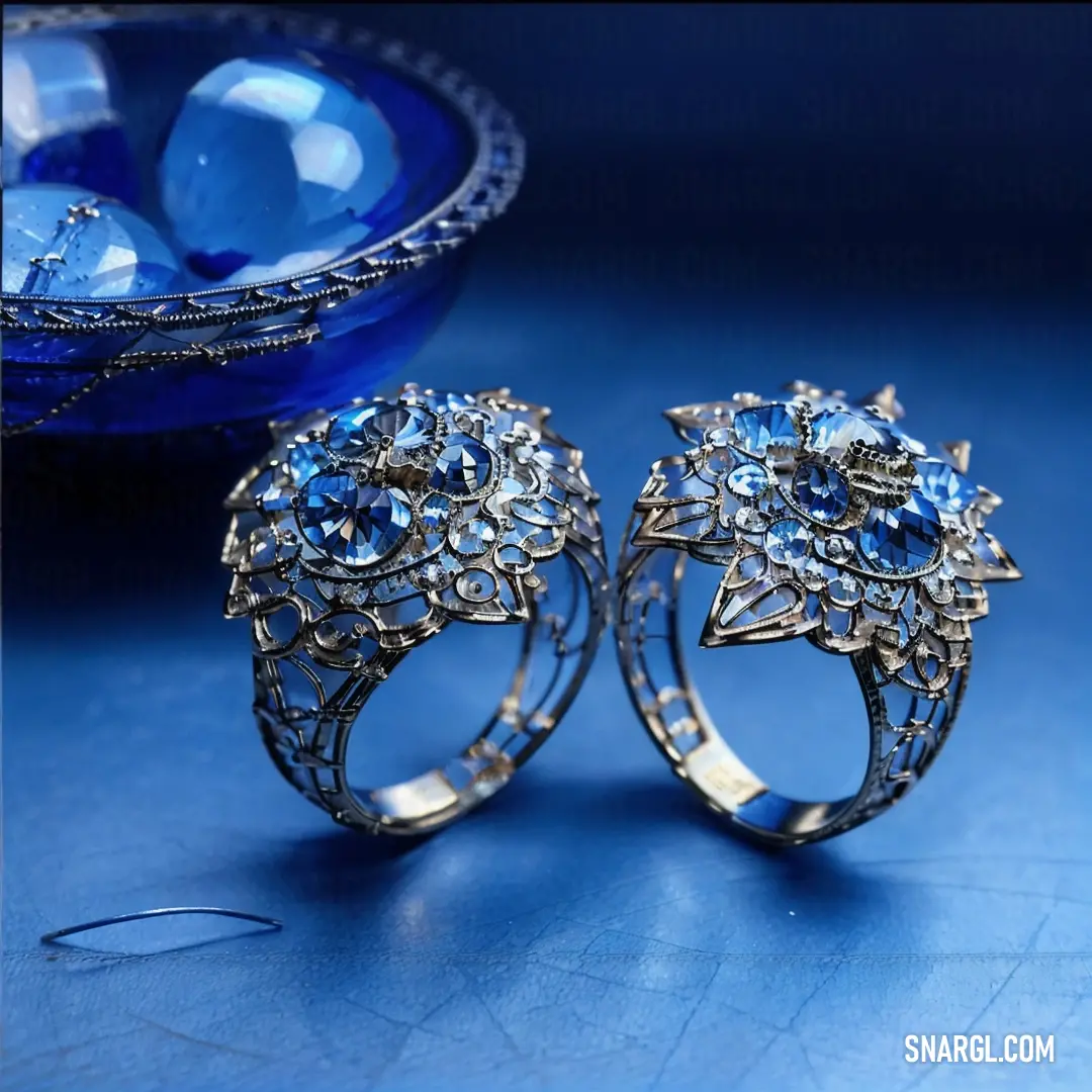 Two rings with blue stones on a blue surface next to a bowl of water