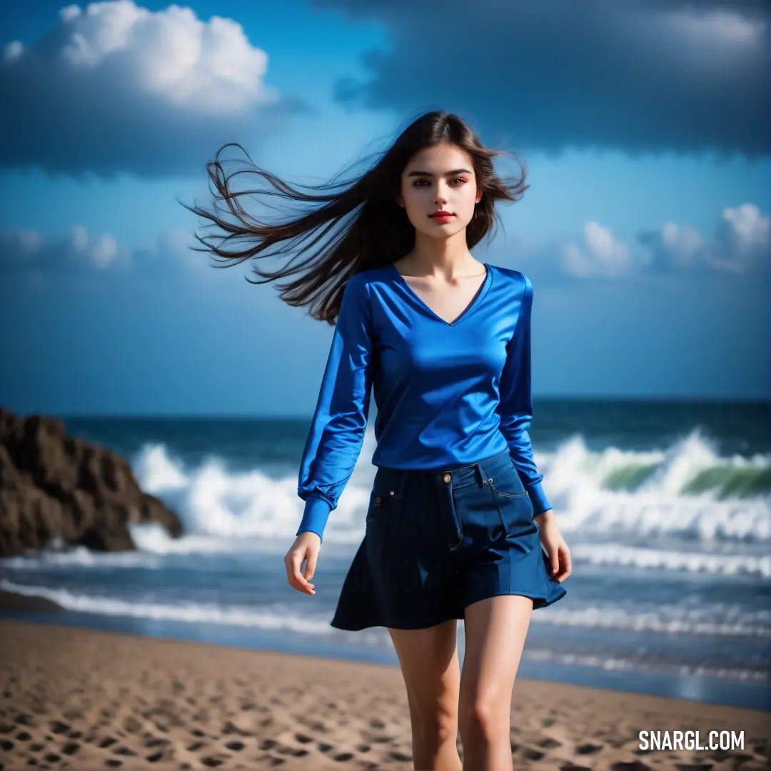 Woman walking on a beach next to the ocean with her hair in the wind and a blue shirt on