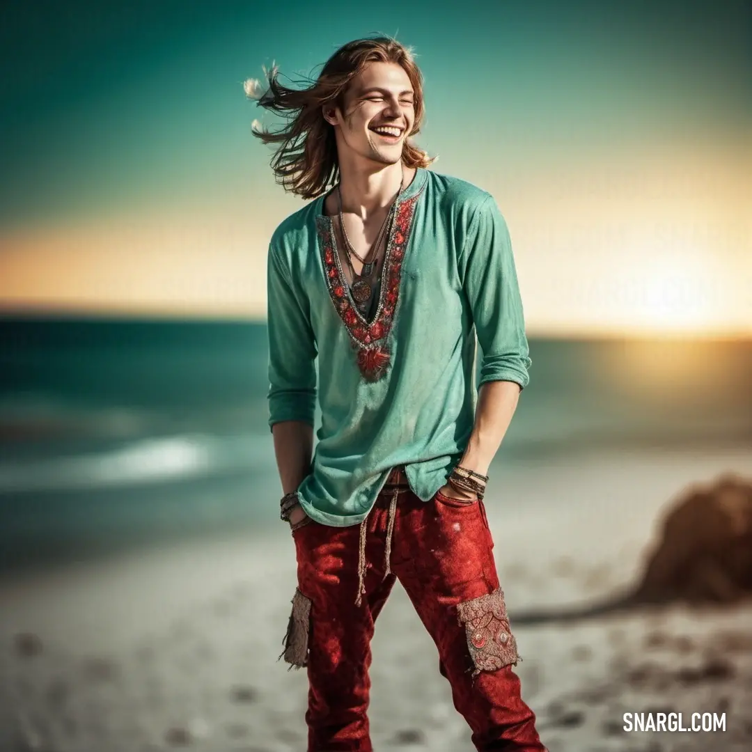 Man with long hair standing on a beach near the ocean with his hands in his pockets and smiling