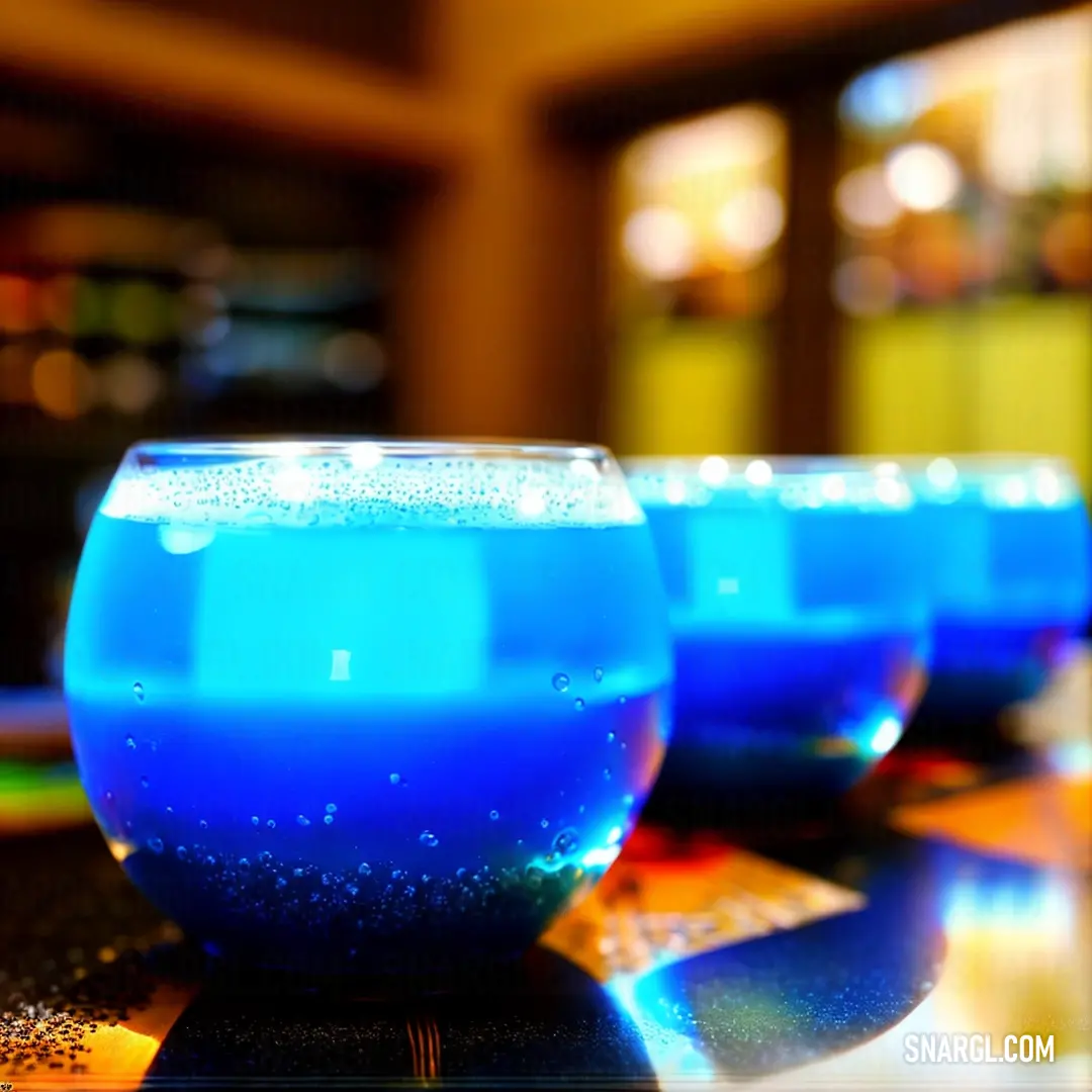 Three glasses filled with a blue liquid, sitting on a table against a blurred background. The vivid blue of the liquid contrasts against the subtle, soft tones of the backdrop, creating a striking visual with a touch of mystery.