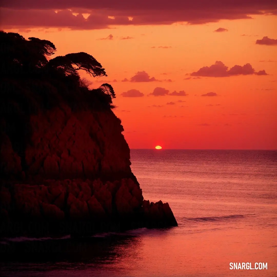 Sunset over the ocean with a large rock outcropping in the foreground and a lone tree on the far side. Example of RGB 207,16,33 color.