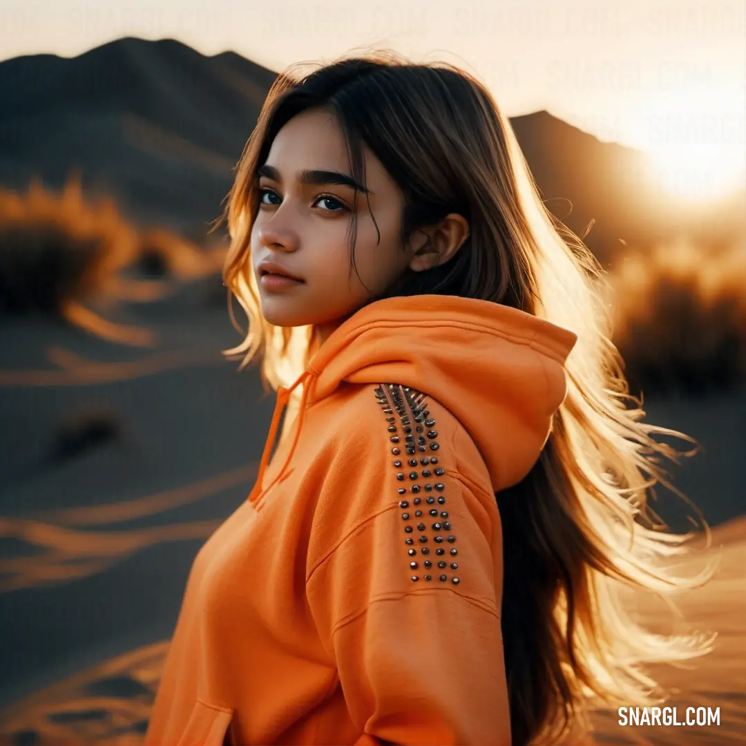 Woman in an orange hoodie standing in the desert at sunset with her hair blowing in the wind. Color #9B4800.