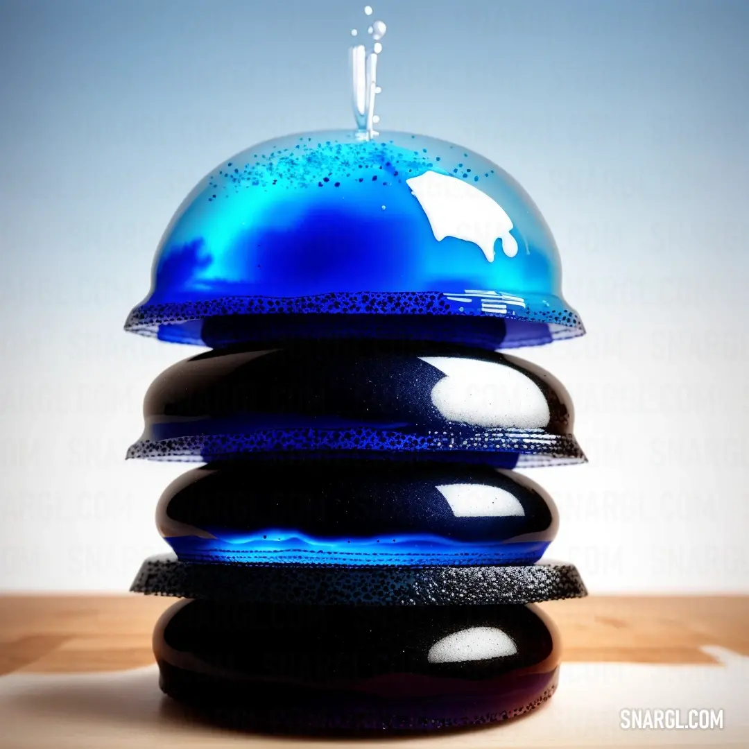 A blue cake cover rests atop a stack of smooth black rocks, the calm blue of the sky serving as the perfect backdrop to the unusual yet serene arrangement. NCS S 6005-Y50R colors tie the scene together, offering a striking contrast.