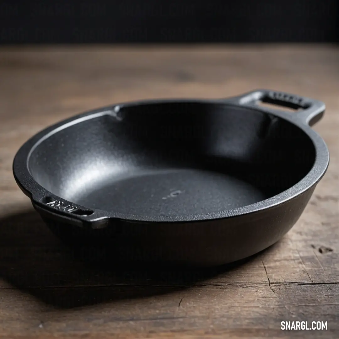 A striking black frying pan rests on a rustic wooden tabletop, showcasing its robust design and complete with a sleek black handle. The captivating colors of CMYK 0,80,30,88 capture the eye, celebrating the beauty of culinary tools in everyday life.