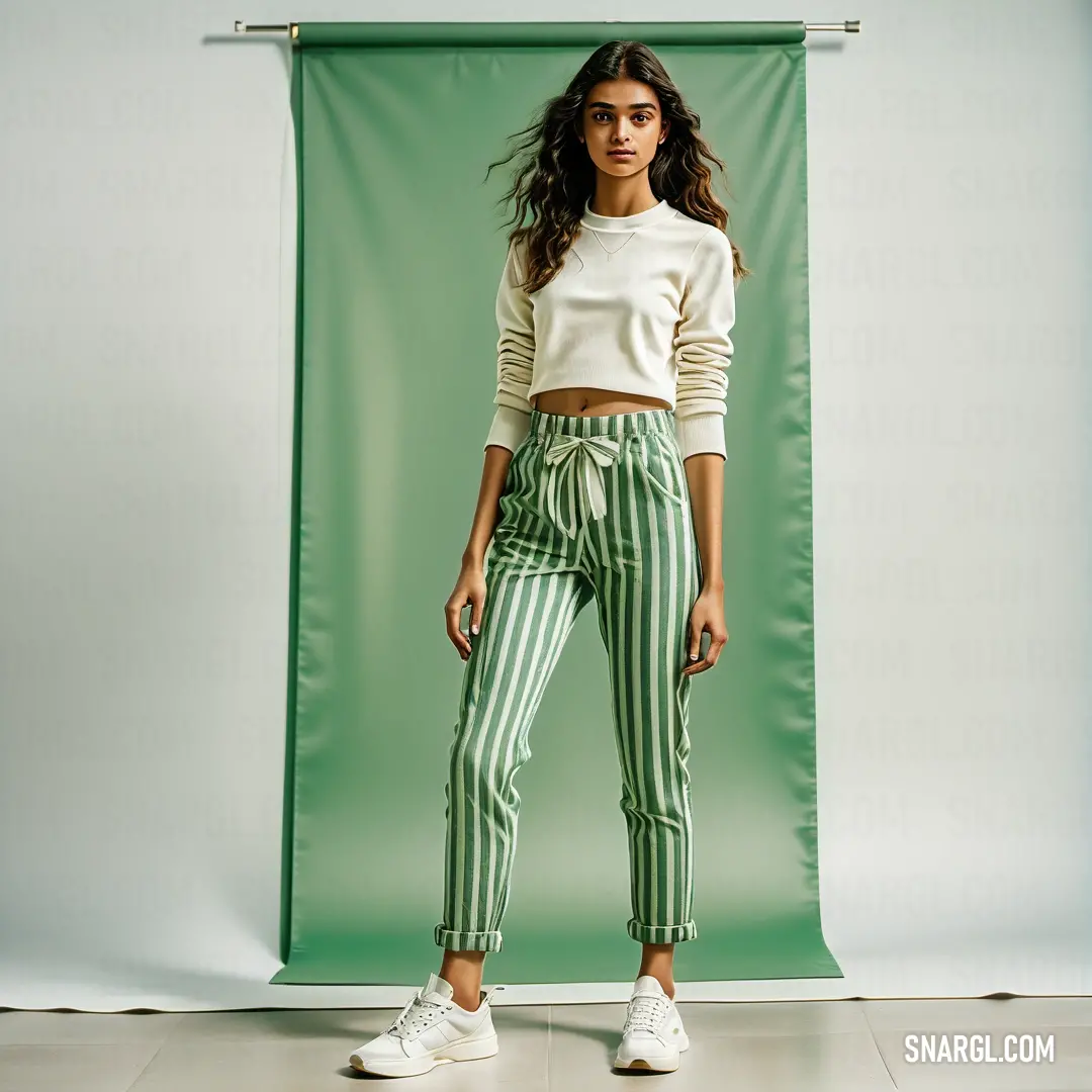 Woman standing in front of a green backdrop wearing a white sweater and green striped pants with a white tennis shoe