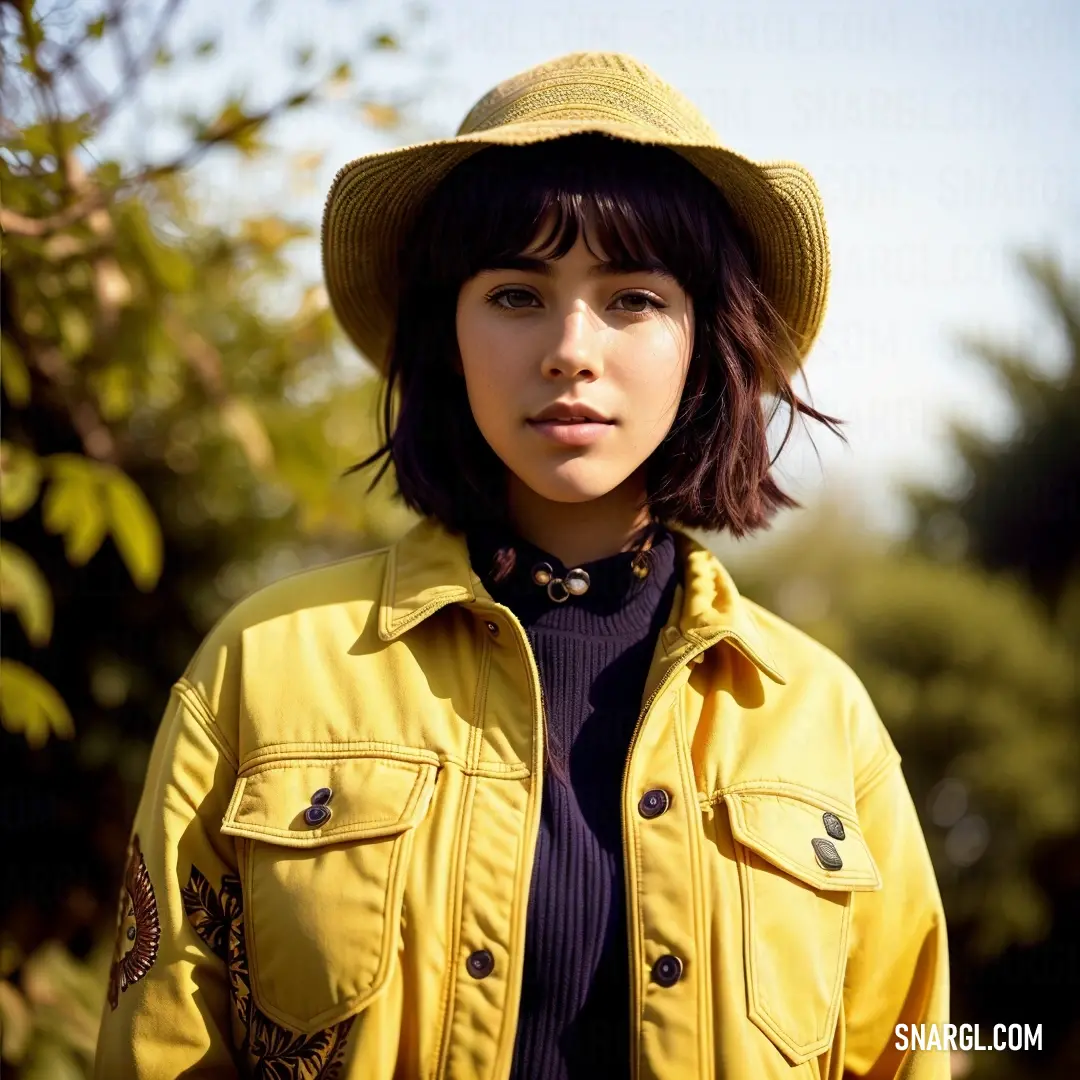 Woman wearing a yellow jacket and hat standing in front of a tree with a blue shirt