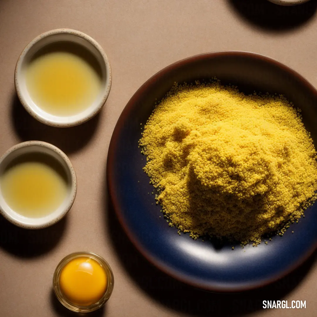 Plate of yellow powder next to three bowls of eggs and a spoon of milk on a table top. Color CMYK 4,53,100,8.