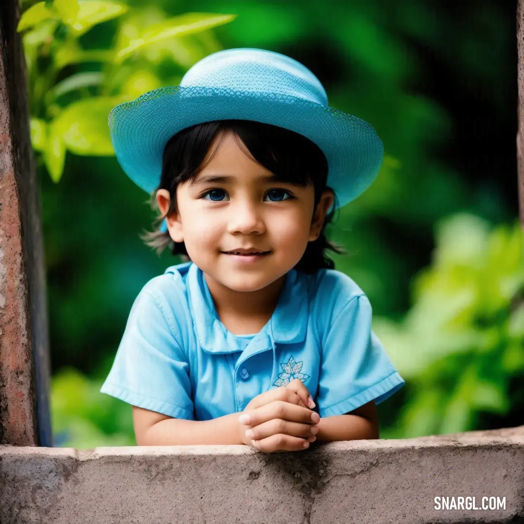 Little girl wearing a blue hat looking out a window with her hands folded on her chest and smiling. Color #4699C5.