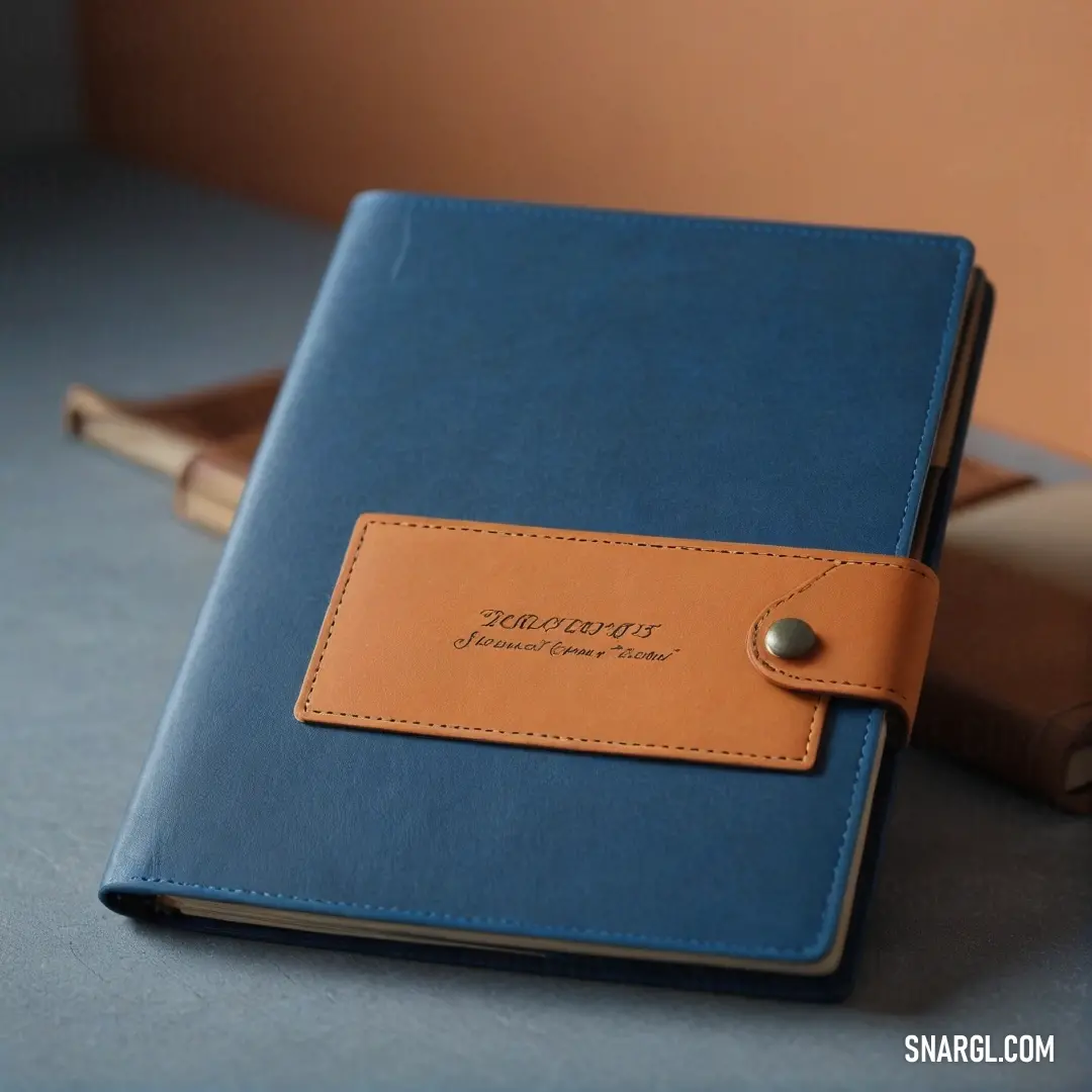 A beautifully crafted blue and tan notebook with a leather cover and a rustic brown strap rests on a table beside an inviting book. The elegant design and warm colors suggest stories waiting to be written and adventures yet to come.