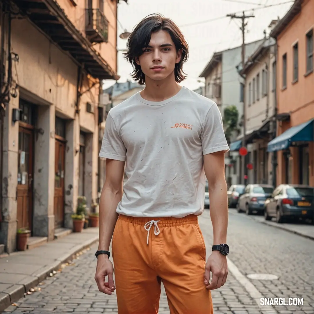 Persian orange color example: Man standing on a street with a white shirt on and orange pants on and a brick road with buildings