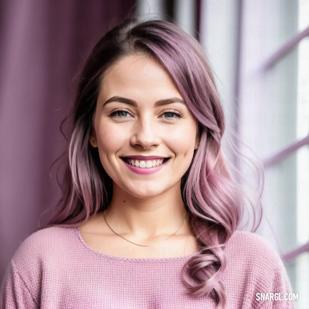 Woman with pink hair smiling at the camera with a window behind her