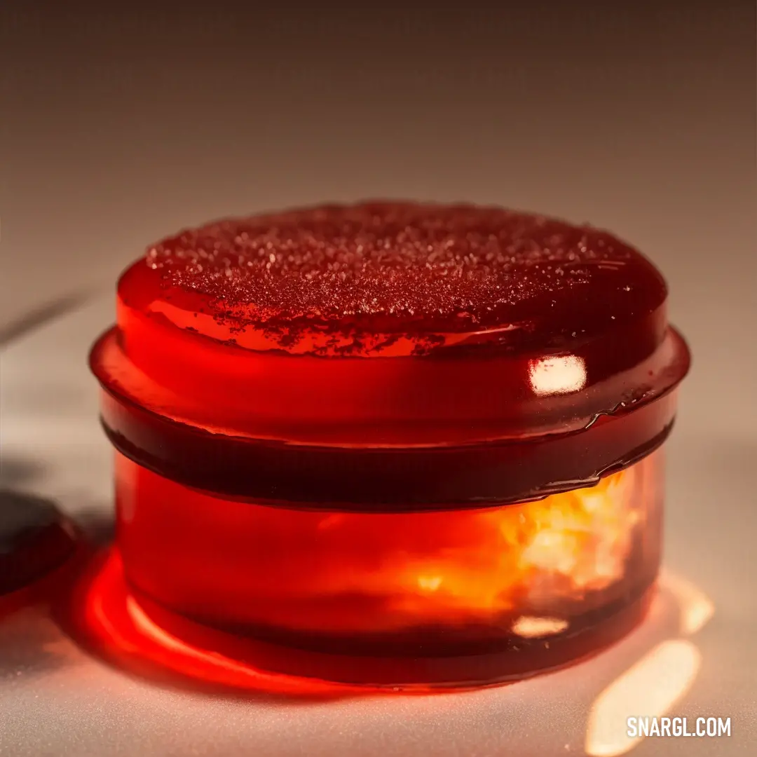 Red jar with a red substance inside of it on a table next to a black object with a white light on it. Example of CMYK 0,100,84,0 color.