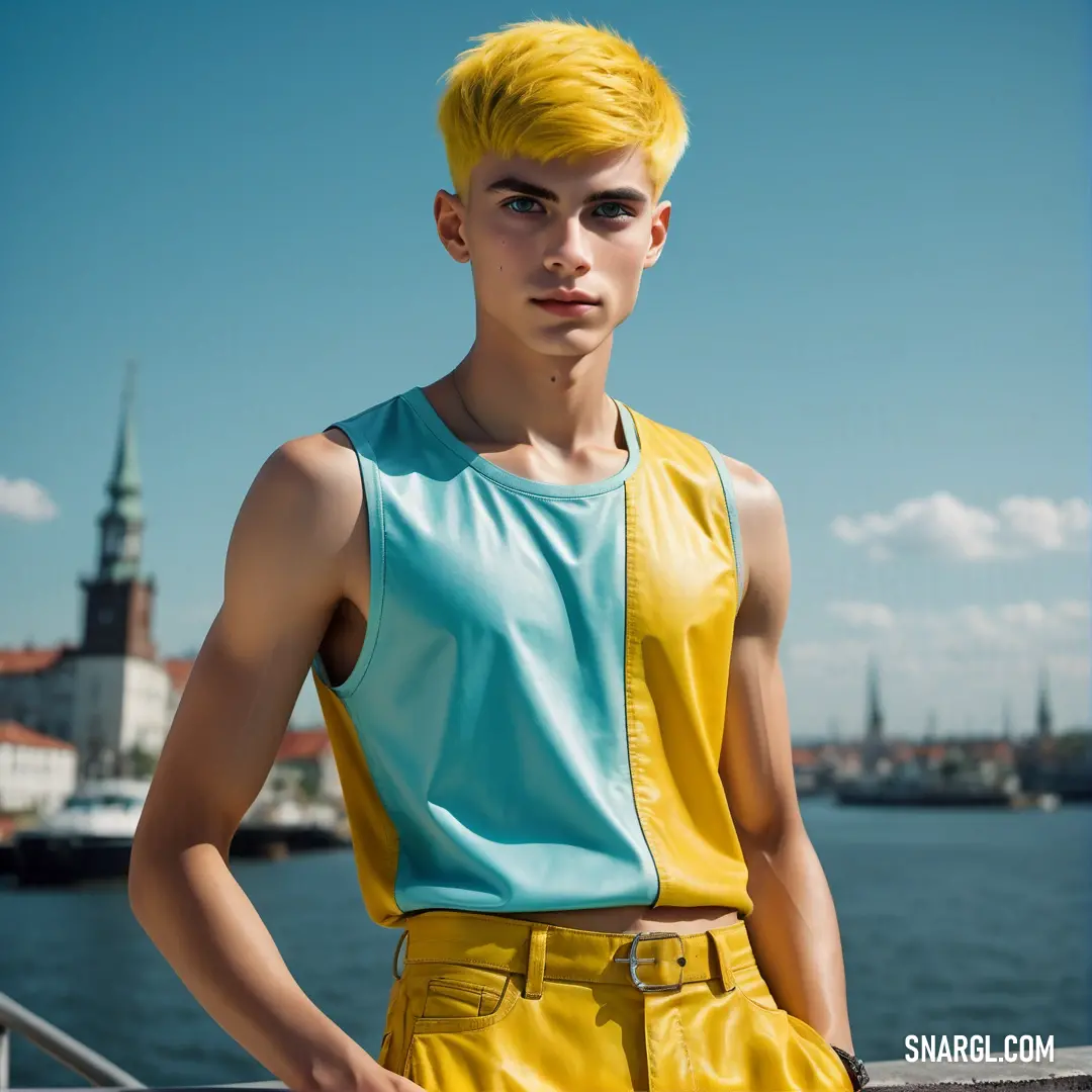 Man with yellow hair and a blue shirt standing on a pier near the water with a city in the background. Example of #ECD540 color.