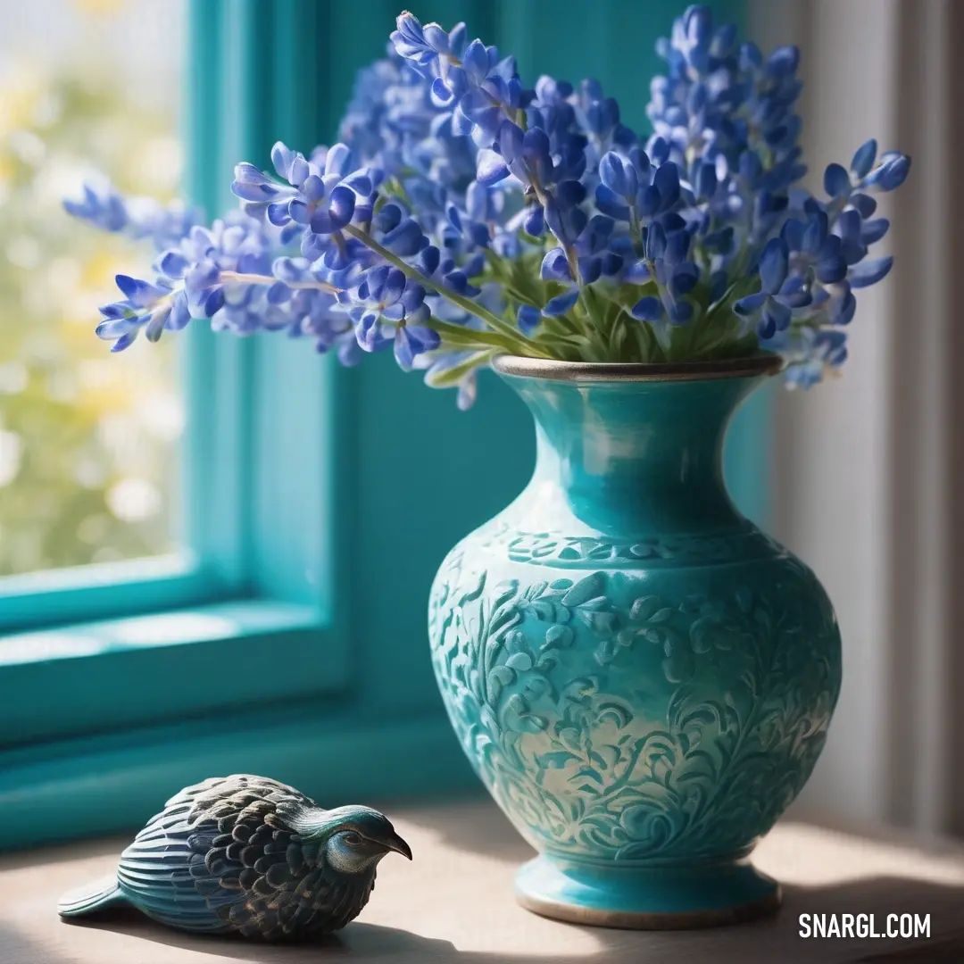 Blue vase with a bird and a flower in it on a table next to a window sill. Color #008080.
