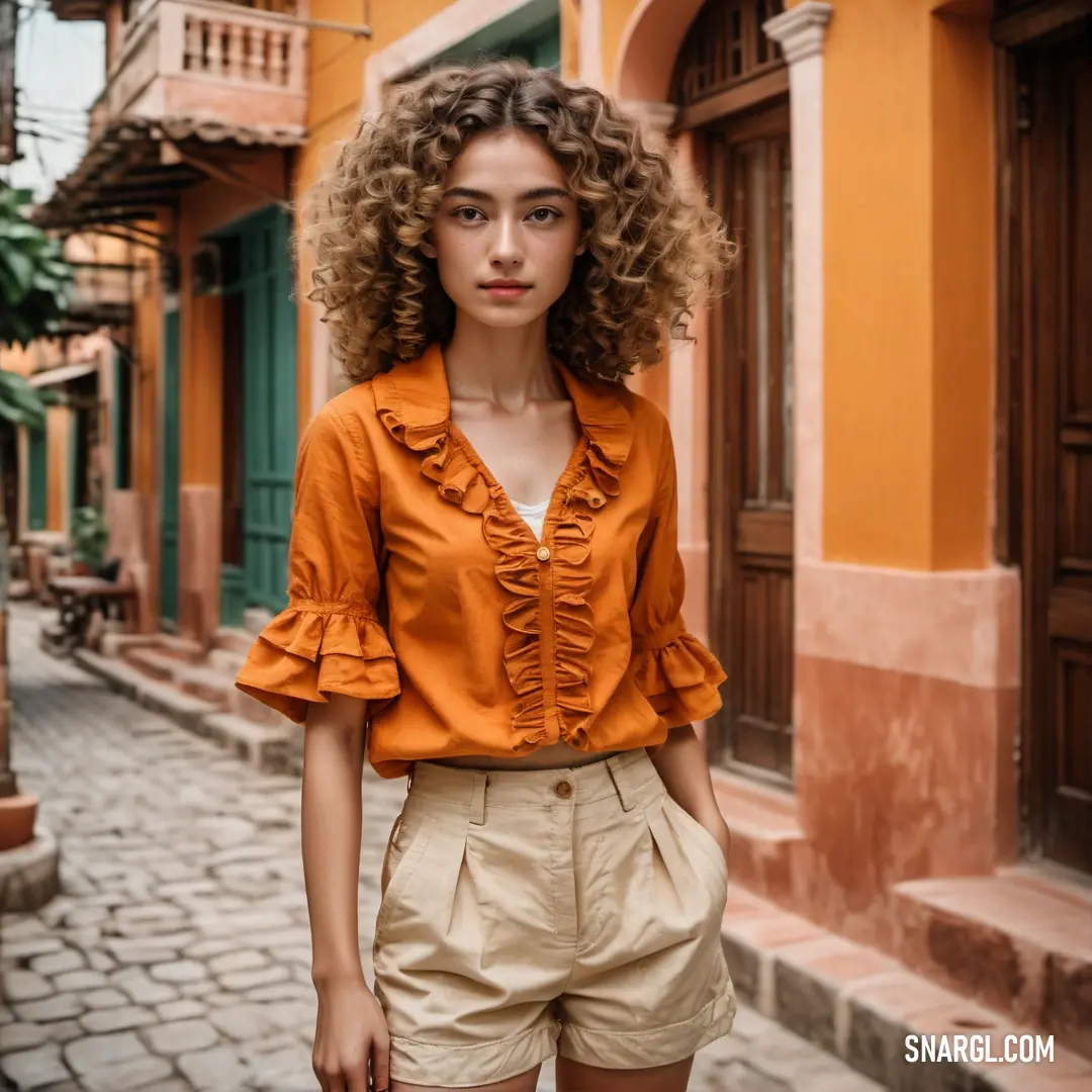 Woman with curly hair standing in front of a building wearing a short skirt and orange shirt with ruffles