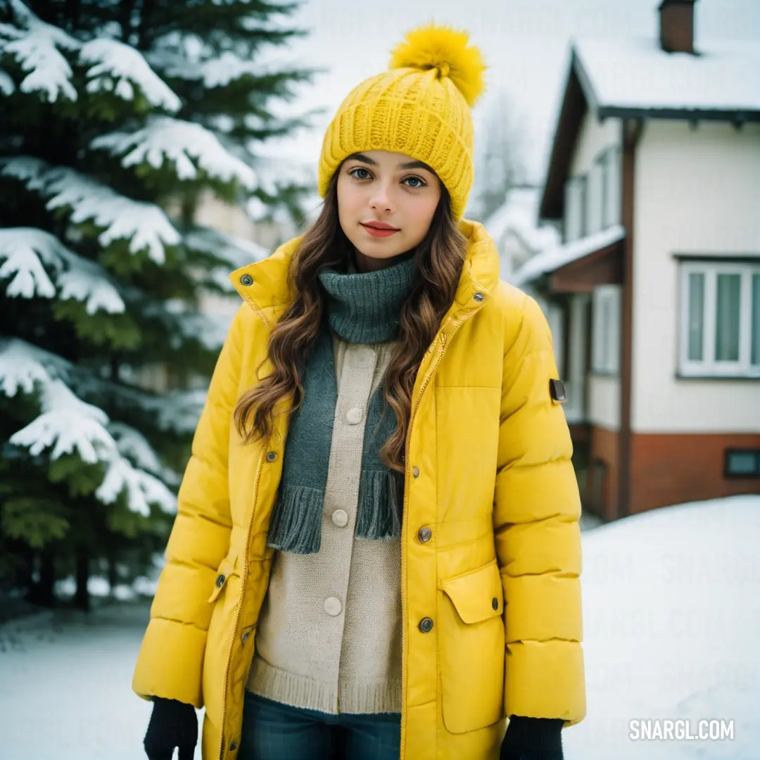 Woman in a yellow jacket and hat standing in the snow in front of a house with a tree